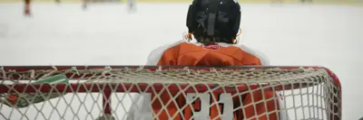 The back view of a young hockey goalie in full gear, standing in front of the net during a game, with the action on the ice blurred in the background.